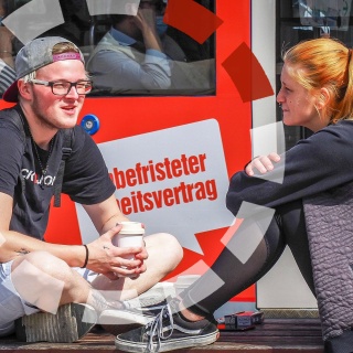 Zwei Jugendliche sitzen vor einer Bahn mit einem Aufkleber: "Befristeter Arbeitsvertrag".