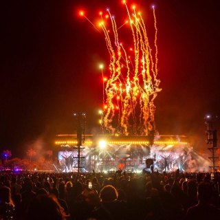 Ein Feuerwerk am Himmel über dem Coachella Valley im US-Bundesstaat Kalifornien. Im Hintergrund ist eine große, hell erleuchtete Bühne mit Musikern zu sehen.