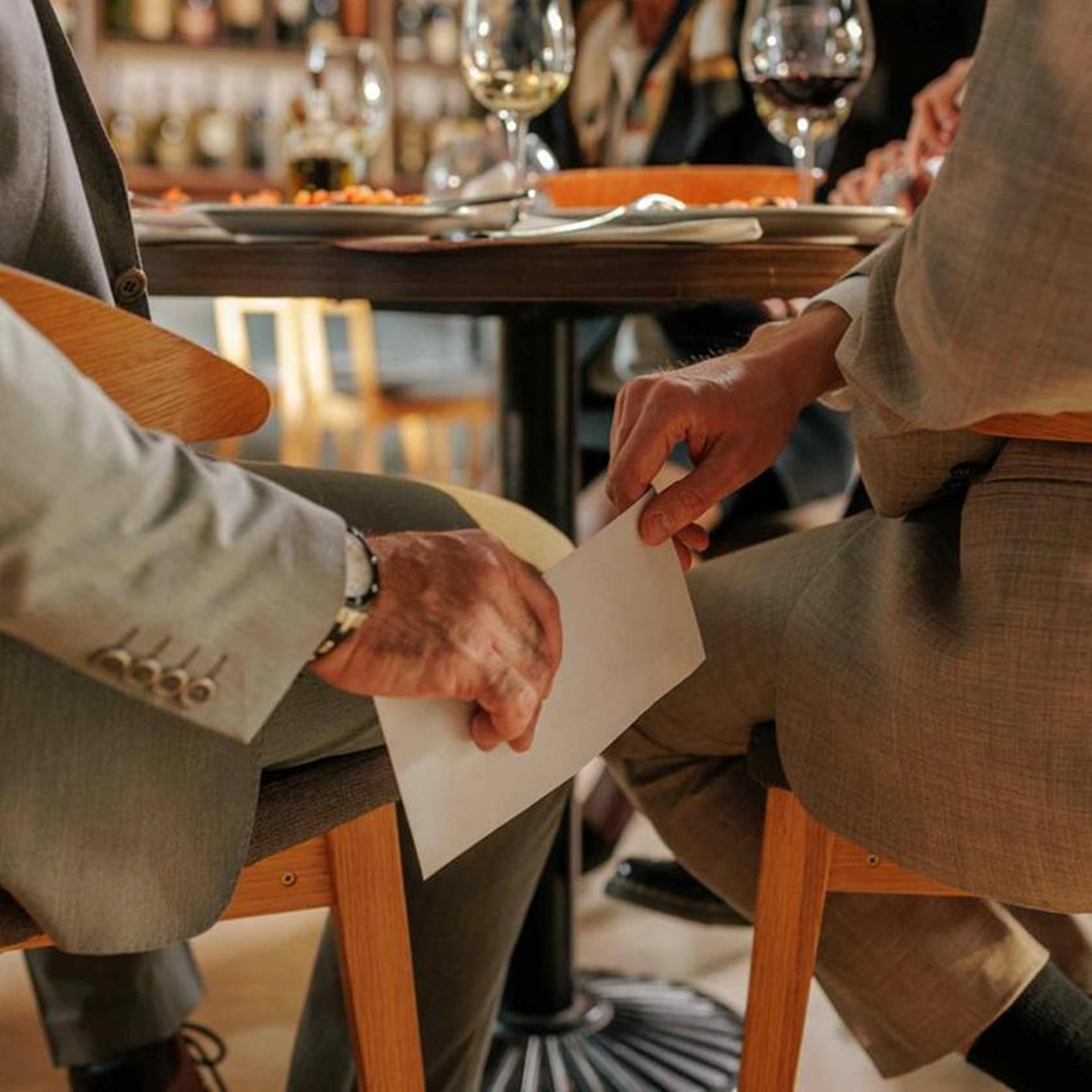In einem Restaurant reicht ein Mann dem anderen unter dem Tisch einen weißen Umschlag.
