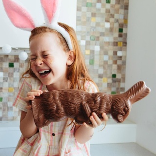 Ein rothaariges Mädchen hält einen Schokoosterhasen in der Hand und ist darüber sichtlich erfreut. Sie trägt einen Haarreifen mit Stoffohren und sieht damit selbst aus wie ein Osterhase.