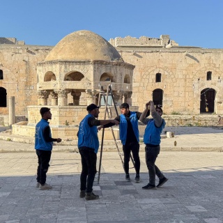 Syrische Arbeiter stehen im Hof einer Moschee und stellen eine Leiter auf. Sie tragen blaue Arbeitswesten.