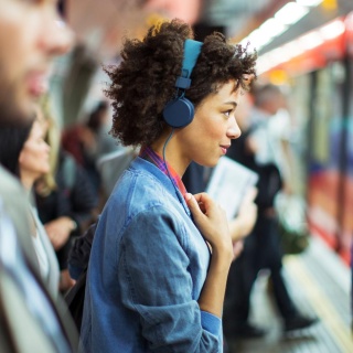 Eine Frau, die im Profil zu sehen ist, trägt blaue Kopfhörer und schaut offen und freundlich geradeaus. Um sie herum sind in der Unschärfe weitere Menschen zu sehen. Sie stehen an einem überfüllten Bahngleis.