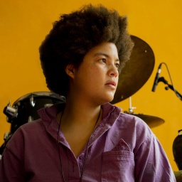 Porträt der Komponistin und Multiinstrumentalistin Natalie Greffel vor einem Drumset, mit leuchtend gelbem Hintergrund.