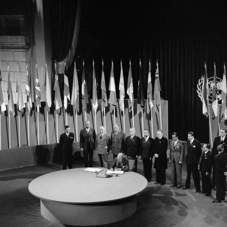 Schwarz-weiss Foto: An einem runden Tisch vor vielen Flaggen und mit zahlreichen Personen im Hintergrund unterschreibt der niederländische Botschafter in den USA die Charta der Vereinten Nationen.