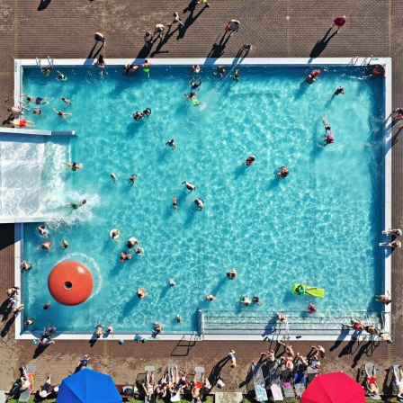 Aufsicht auf ein Badebecken in einem Freibad. Einige Leute schwimmen, andere vergnügen sich auf der großen Rutsche.