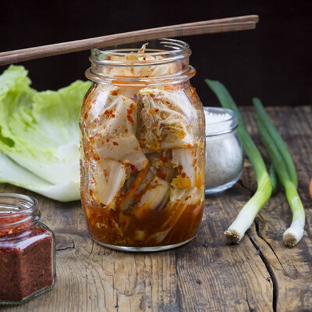Ein Glas Kimchi, Kohl, Frühlingszwiebeln und eine Karotte auf einem Holztisch.