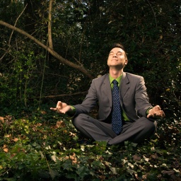 Young businessman sits in a lotus position meditating in the woods with closed eyes and a smile.