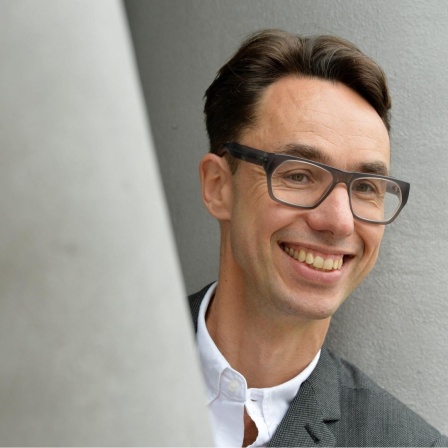 Hinter einem grauen Pfeiler schaut ein Mann mit dunklen zurückgekämmten Haaren und großer Brille und einem freundlichen Gesicht hervor. Er trägt ein weißes Hemd und eine Anzugjacke.