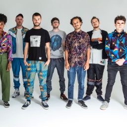 Die zehn Mitglieder der multi-ethnischen britischen Bigband Nubiyan Twist vor weißem Hintergrund
