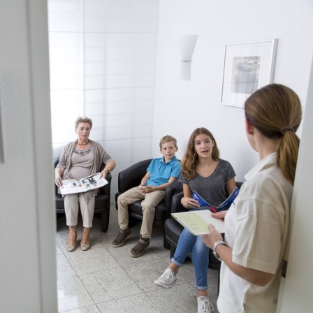 Sprechstundenhilfe ruft in einem Wartezimmer einen Patienten auf.