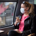 Frau mit Mundschutzmaske, sitzt in Zug, am Handy, Corona-Krise