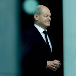 Olaf Scholz trägt einen schwarzen Anzug und steht zwischen zwei grauen Säulen, die in der Unschärfe zu erkennen sind