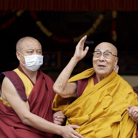 Der Dalai Lama winkt während einer Gebetszeremonie, während er von zwei Mönchen gestützt wird.