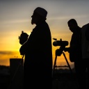 Die Silhouetten syrischer Gelehrter, die mit einem Teleskop den Mond beobachten, zeichnen sich gegen den rötlich schimmernden Himmel ab.