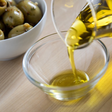 Olivenöl wird in eine Glasschale gefüllt während im Hintergrund eine Schale mit grünen Oliven steht.