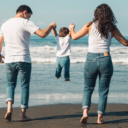 Ein Elternpaar hält sein kleines Kind am Strand an den Händen und lässt es fliegen