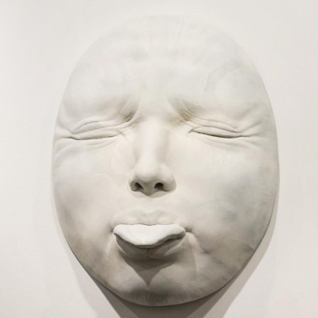 Der spanische Künstler Samuel Salcedo vor der, von ihm geschaffenen Plastik eines hyperrealistischen Gesichts, mit herausgestreckter Zunge.