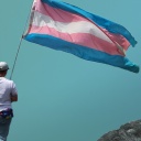 Dean steht in Jeans und weißem T-Shirt von hinten Fotografiert mit einer blau-rosa-weiß gestreiften Transflagge in der Hand vor einem blauen HIntergrund mit Bergen.