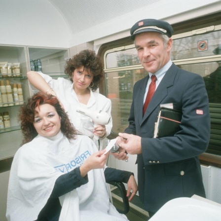 Intercity-Friseur im Mai 1989. Eine Frau bekommt die Haare frisiert, ein Zugbegleiter kontrolloiert den Fahrschein: