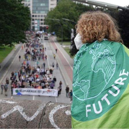 Ein Mensch mit einer Fahne mit dem Logo von Fridays for Future verfolgt von einer Brücke aus die Demonstration des Bündnisses Fridays for Future entlang einer Straße. 