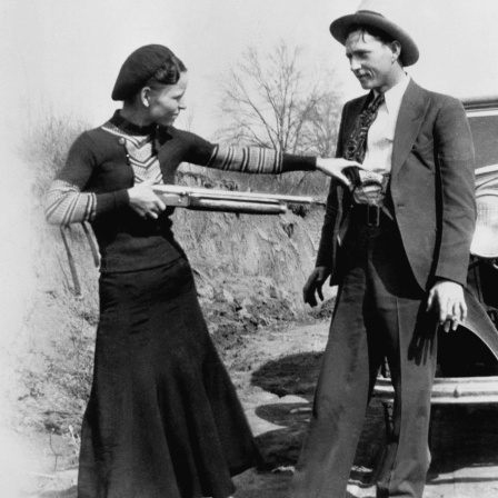 Bonnie Parker in dunklem Kleid und Clyde Barrow im Anzug, posieren mit einer Flinte vor einem Auto.
