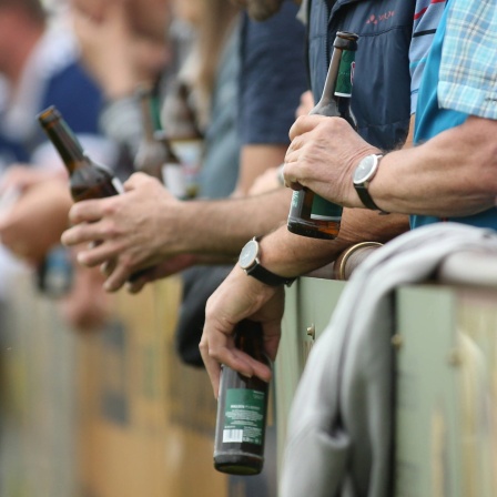 Männer stehen mit Bierflasche hinter einer Bande.