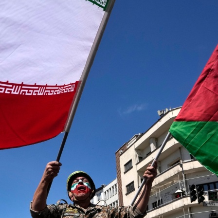 Ein Mann, dessen Gesicht in den Farben der iranischen Fahne bemalt ist, schwenkt links die iranische und rechts die palästinensische Fahne.