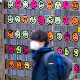 Eine Passantin mit Mund-Nasen-Bedeckung geht an einem hölzernen Bauzaun vorbei, der mit bunten Smileys bemalt wurde (Bild: dpa / Peter Kneffel)