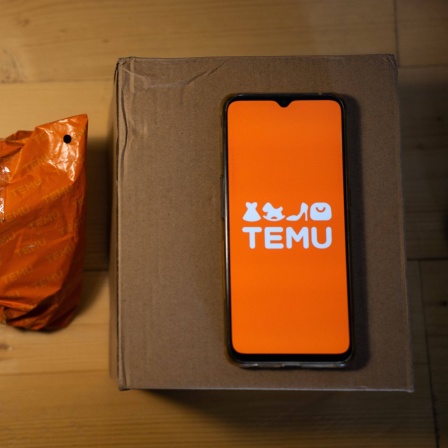 Auf einem Paket liegt ein Smartphone das die App von Temu zeigt, daneben eine orangene Versandtasche von Temu