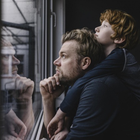 Ein Vater schaut mit seinem Sohn auf dem Rücken nachdenklich aus dem Fenster.