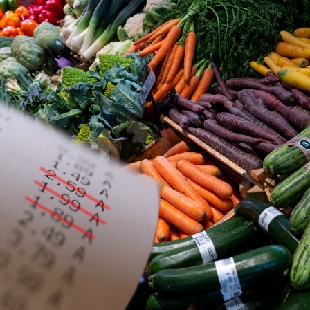 Ein Kassenzettel mit durchgestrichenen Preisen in einer Detailaufnahme, neben dem gut gefüllten Gemüseregal eines Supermarkts.