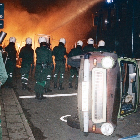 Polizeibeamte bekaempfen am Abend des 24. August 1992 in Rostock-Lichtenhagen eine brennende Strassenblockade mit einem Wasserwerfer. Im Vordergrund liegt ein Auto auf der Seite