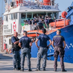 Auf einem Schiff im Hafen von Lampedusa warten viele Menschen darauf, an Land gehen zu können. Vor dem Schiff stehen fünf Polizisten.