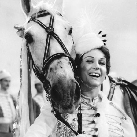 Marika Rökk trägt eine ungarische Tracht und lehnt lachend an einem Pferd. (Filmstill)