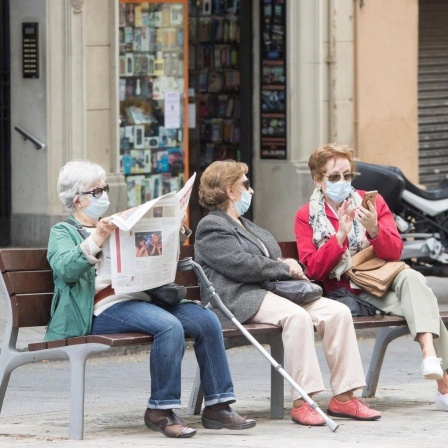 Drei ältere Frauen sitzen in mit Coronamaske in Barcelona auf einer Bank. Eine Frau liest Zeitung, die beiden anderen unterhalten sich. 