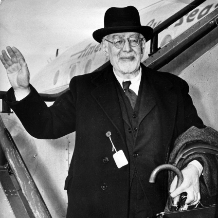 Eine Schwarzweiß-Fotografie zeigt einen älteren Mann mit schwarzem Hut, grüßend die Rolltreppe eines Flugzeugs herabsteigend