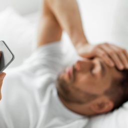 Ein im Bett liegender Mann hält sich mit einer Hand den Kopf, in der anderen ein Smartphone