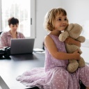 Eine Frau arbeitet an einem Tisch mit dem Laptop. Auf dem Tisch sitzt ein Mädchen, das einen Teddybären im Arm hält.