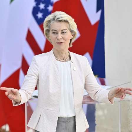 Ursula Von der Leyen mit ausgebreiteten Armen vor den Flaggen der G7-Staaten