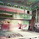 Das Bild zeigt ein Farbfoto der Künstlerin Johanna Diehl aus ihrer Serie "Ukraine Series". Zu sehen ist ein halb verfallener Saal mit altrosa Wänden, von denen die Farbe abblättert. Die Decke ist mit kleinen Gemälden geschmückt
      