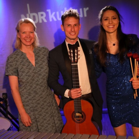 In der Mitte steht ein junger Mann mit einer Gitarre in der Hand und rechts und links neben ihm eine Frau.