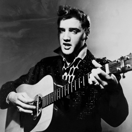 Elvis Presley mit Gitarre vor grauem Hintergrund MItte der 1950er Jahre.