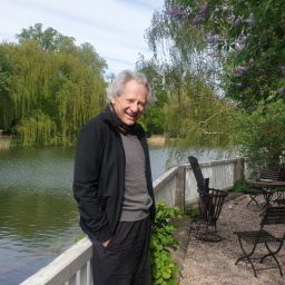 Ein lachender Mann lehnt an der Brüstung eines Zauns vor einem Teich in einem Park.