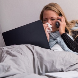 Eine Frau liegt krank im Bett. Telefoniert mit einem Laptop auf den Knien und putzt sich die Nase