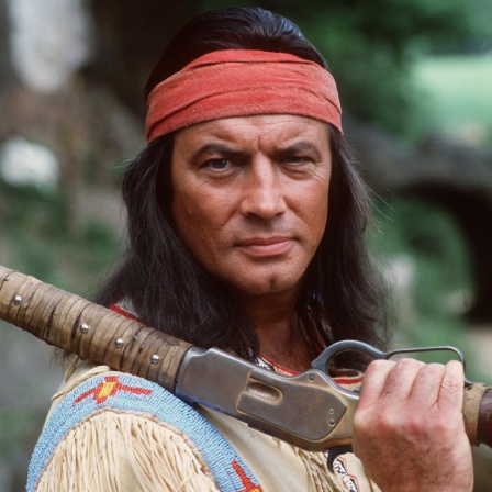 Pierre Brice als Winnetou. Er trägt ein Wildlederkostüm mit Fransen, ein rotes Stirntuch und ein Gewehr über der Schulter.