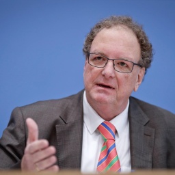 Älterer Mann mit hoher Stirm und angegrautem gelocktem Haar sitzt im Anzug vor einer blauen Wand und hebt eine Hand geradeaus vor seinen Oberkörper.