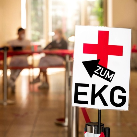 "Zum EKG!" steht auf einem Schild einer Arztpraxis - im Hintergrund zu sehen sind medizinische Fachangestellte. (Symbolbild) 