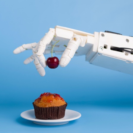 Eine Roboterhand setzt eine Kirsche auf einen kleinen Muffin. 