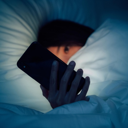 Zu sehen ist eine Frau in der Unschäfe, eingehüllt in eine dicke Bettdecke. Sie blickt auf ein leuchtendes Smartphone, dass ihr besorgtes Gesicht im Dunkeln erhellt.
