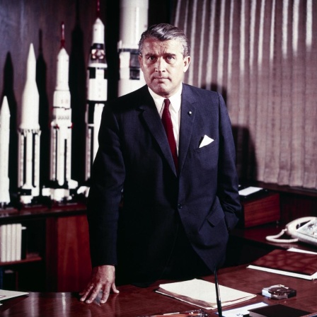 Wernher von Braun steht die Hand auf dem Schreibtisch gestützt in seinem Büro. Im Hintergrund sind Raketenmodelle zu sehen.